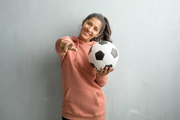 Young sporty indian woman against a wall cheerful and smiling pointing to the front. Holding a soccer ball to play a game.