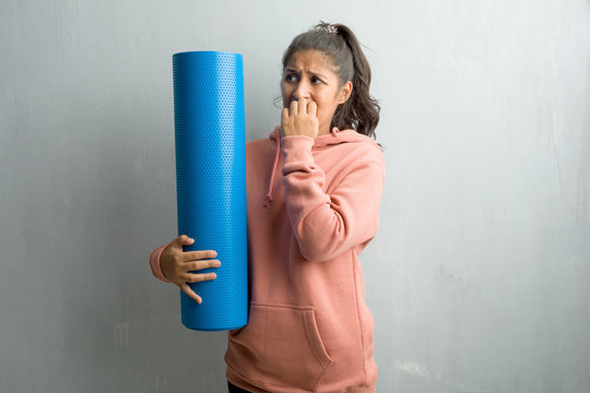 Young Sporty Indian Woman Against A Wall Biting Nails, Nervous And Very Anxious And Scared For The Future, Feels Panic And Stress. Holding A Blue Mat To Practice Yoga.