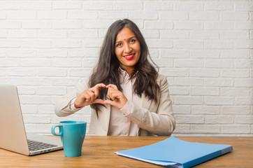 Young indian woman at the office making a heart with hands, expressing the concept of love and friendship, happy and smiling