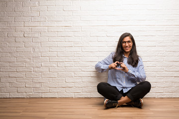 Young indian woman sit against a brick wall making a heart with hands, expressing the concept of love and friendship, happy and smiling