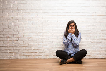 Young indian woman sit against a brick wall biting nails, nervous and very anxious and scared for...