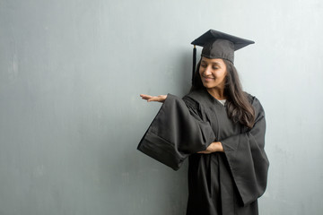 Young graduated indian woman against a wall holding something with hands, showing a product, smiling and cheerful, offering an imaginary object