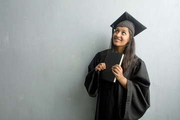 Young graduated indian woman against a wall looking up, thinking of something fun and having an idea, concept of imagination, happy and excited. Holding a book.