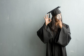 Young graduated indian woman against a wall cheerful and confident doing ok gesture, excited and screaming, concept of approval and success. Holding a magnifying glass.