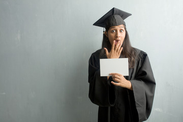 Young graduated indian woman against a wall surprised and shocked, looking with wide eyes, excited by an offer or by a new job, win concept. Holding a placard.
