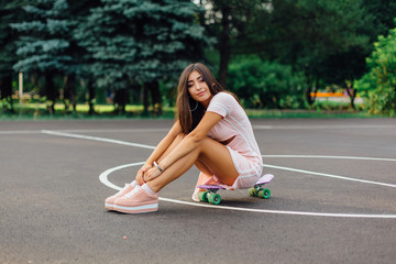 Portrait of a smiling charming brunette female sitting on her skateboard on a basketball court.