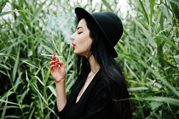 Sensual smoker girl all in black, red lips and hat. Goth dramatic woman smoking on common reed.