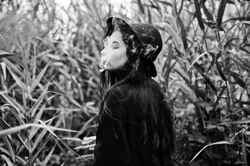 Sensual smoker girl all in black, red lips and hat. Goth dramatic woman smoking on common reed....