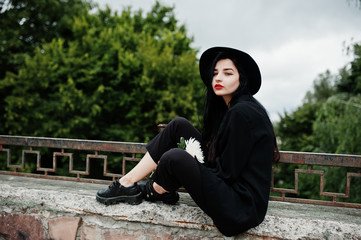 Sensual girl all in black, red lips and hat. Goth dramatic woman hold white chrysanthemum flower.