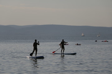 Two men floating on boards. Stand up paddle on Lake Balaton. Beautiful, warm, sunny, calm summer day. Mountains, sailboats, blue sky visible in the background. Active rest during the holidays.