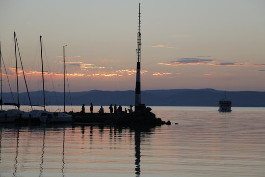 Ship sailing to the port. A warm, pleasant summer evening. Twilight on Lake Balaton. A quiet end of the day. Reflection on the surface of the water. Mast on the breakwater. Mountains in the background