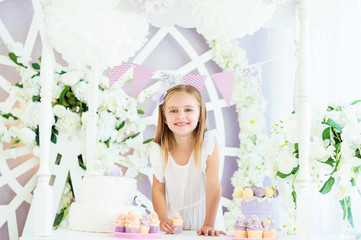 Obraz na płótnie Canvas Pretty little smiling girl standing at the white table with cakes in the beatiful decorated candy bar