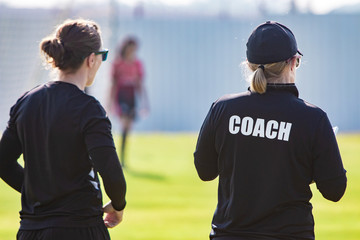 Back view of female sport coach and her assistant in black COACH shirt at an outdoor sport field - obrazy, fototapety, plakaty