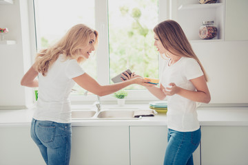 Close up side profile photo two people mum and teen daughter house work duties deny wash plates want use user telephone problem trouble rage wear white t-shirts jeans in bright flat kitchen