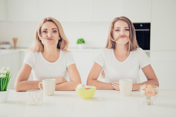 Close up photo two people mum teen daughter pretending man male guy making moustache with hair funny funky fooling around beverage sweets on table wear white t-shirts jeans in bright flat kitchen