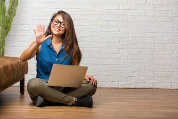 Portrait of young latin woman sitting on the floor showing number five, symbol of counting, concept of mathematics, confident and cheerful. Holding a laptop.