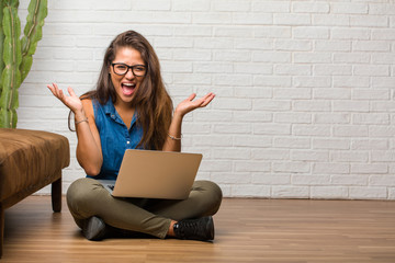 Portrait of young latin woman sitting on the floor screaming happy, surprised by an offer or a promotion, gaping, jumping and proud. Holding a laptop.