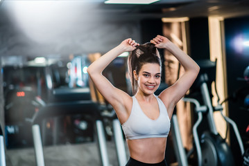Fototapeta na wymiar Ready to workout! Beautiful smiling girl in sportswear tying her hair while standing in the gym, looking at camera.