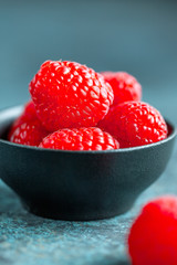 Fresh ripe raspberries in a small black bowl on a textured blue table. Macro photography.