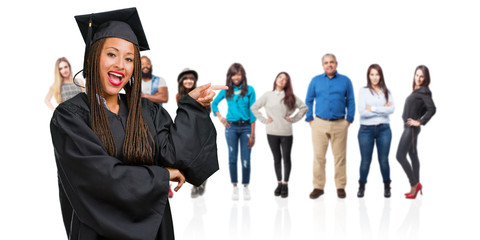 Young graduated black woman wearing braids pointing to the side, smiling surprised presenting something, natural and casual