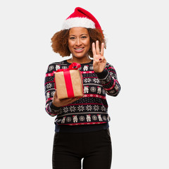 Young black woman holding a gift in christmas day showing number three