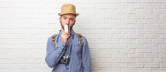 Young traveler man wearing backpack and a vintage camera keeping a secret or asking for silence, serious face, obedience concept. Holding a credit card.