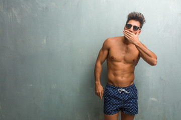 Young athletic man wearing a swimsuit against a grunge wall covering mouth, symbol of silence and repression, trying not to say anything