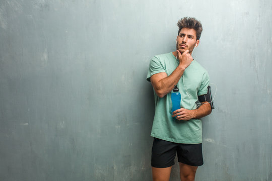 Young fitness man against a grunge wall thinking and looking up, confused about an idea, would be trying to find a solution. Holding a blue energy drink.