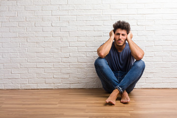 Young natural man sit on a wooden floor covering ears with hands, angry and tired of hearing some sound