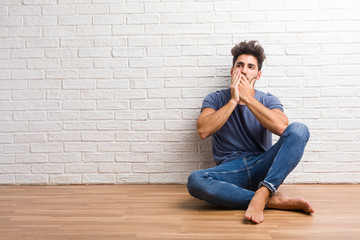Young natural man sit on a wooden floor covering mouth, symbol of silence and repression, trying not to say anything