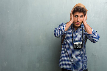 Young handsome traveler man wearing a straw hat, a backpack and a photo camera covering ears with hands, angry and tired of hearing some sound