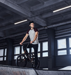 Fototapeta na wymiar Full body portrait of a young man in protective helmet with his bike standing in a skatepark indoors