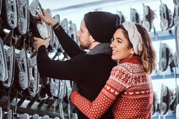 Young attractive couple wearing warm clothes standing together, choosing pairs of skates near rack inside.