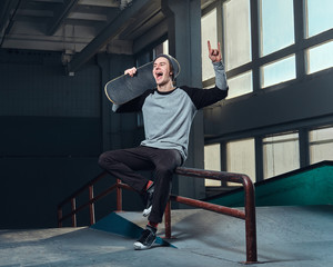 Cheerful young man wearing a shirt and hat holding his board, sitting on a grind rail in skatepark indoors.