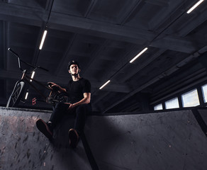 Young Bmx rider relaxing after practicing tricks with his bike in a skatepark indoors