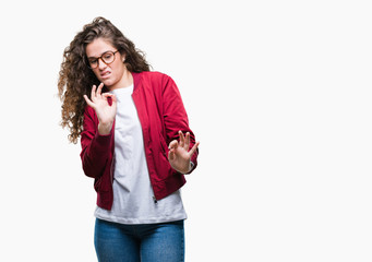 Beautiful brunette curly hair young girl wearing jacket and glasses over isolated background disgusted expression, displeased and fearful doing disgust face because aversion reaction