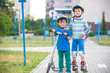 Two kid boy on roller skates and his sibling brother on scooter wrapped in park. Children wearing...