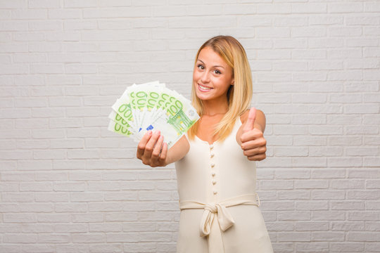 Portrait of young pretty blonde woman against a bricks wall cheerful and excited, smiling and raising her thumb up, concept of success and approval, ok gesture. Holding euro banknotes.