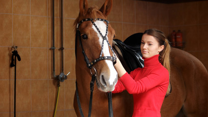 Beautiful girl with her horse in stable. Young woman adjusting her horse bridle before a ride. People and animals concept. Equine business.