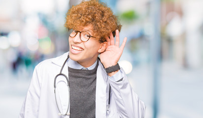 Young handsome doctor man wearing medical coat smiling with hand over ear listening an hearing to rumor or gossip. Deafness concept.