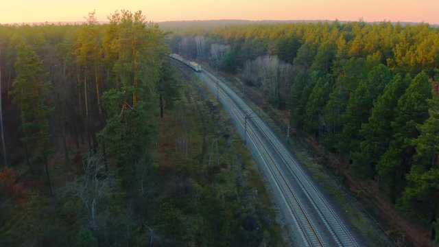 Aerial oncoming shot of a commuter train moving through the autumn forest at sunset
