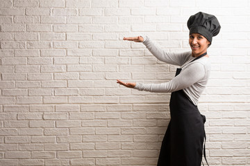 Young baker indian woman against a bricks wall holding something with hands, showing a product, smiling and cheerful, offering an imaginary object