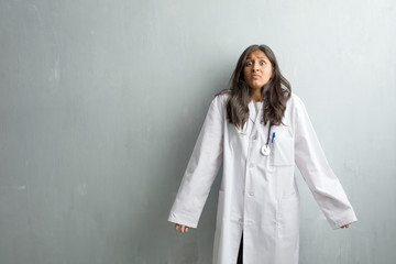 Young indian doctor woman against a wall doubting and shrugging shoulders, concept of indecision and insecurity, uncertain about something - obrazy, fototapety, plakaty