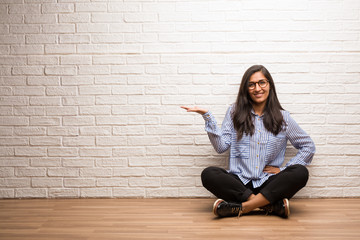 Young indian woman sit against a brick wall holding something with hands, showing a product, smiling and cheerful, offering an imaginary object