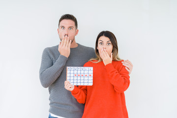 Young couple holding ovulation calendar over isolated background cover mouth with hand shocked with...