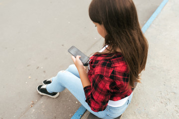 Girl teenager 11-12 years old sits on skateboard in park summer against background concrete stairs. In hands smartphone writes message in Internet application. Teenagers in social networks. Back view.