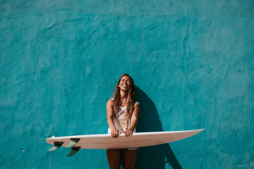 happy surfer girl with surfboard in front of blue wall - obrazy, fototapety, plakaty