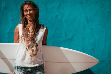 happy surfer girl with surfboard in front of blue wall