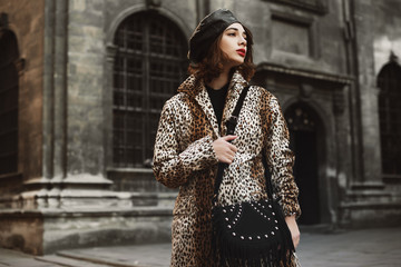 Outdoor fashion portrait of young woman wearing trendy leopard print coat, leather beret, holding...