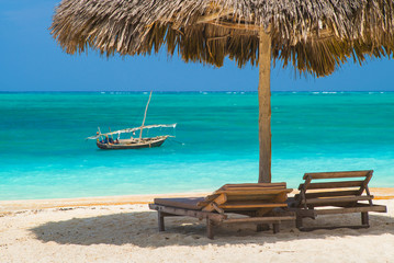 sun chairs under wooden beach umbrella with view to emerald lagoon with boat in Zanzibar in Tanzania